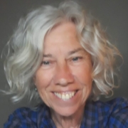 Smiling older woman with curly grey hair, wearing a blue checked shirt, in front of a plain background.