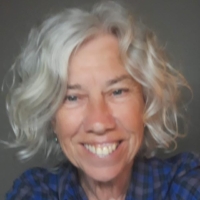 Smiling older woman with curly grey hair, wearing a blue checked shirt, in front of a plain background.