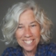 Smiling older woman with curly grey hair, wearing a blue checked shirt, in front of a plain background.