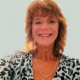 Woman with shoulder-length reddish hair, wearing a leopard print top and a rose pendant necklace, smiling.