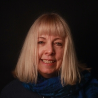 Smiling woman with straight blonde hair and fringe, wearing a blue scarf, in front of a dark background.