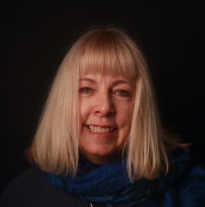 Smiling woman with straight blonde hair and fringe, wearing a blue scarf, in front of a dark background.
