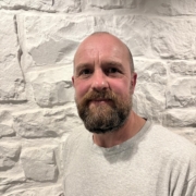 A bearded man in a grey T-shirt stands smiling in front of a textured white stone wall.