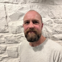 A bearded man in a grey T-shirt stands smiling in front of a textured white stone wall.