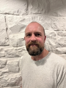 A bearded man in a grey T-shirt stands smiling in front of a textured white stone wall.