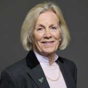 Older woman with short blonde hair in a suit, smiling against a plain dark background.