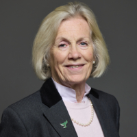 Older woman with short blonde hair in a suit, smiling against a plain dark background.