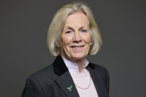 Older woman with short blonde hair in a suit, smiling against a plain dark background.