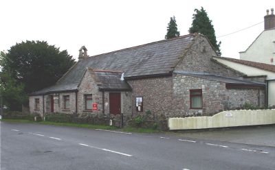Stone building with small windows and pitched roof beside a road, surrounded by trees and a low fence.